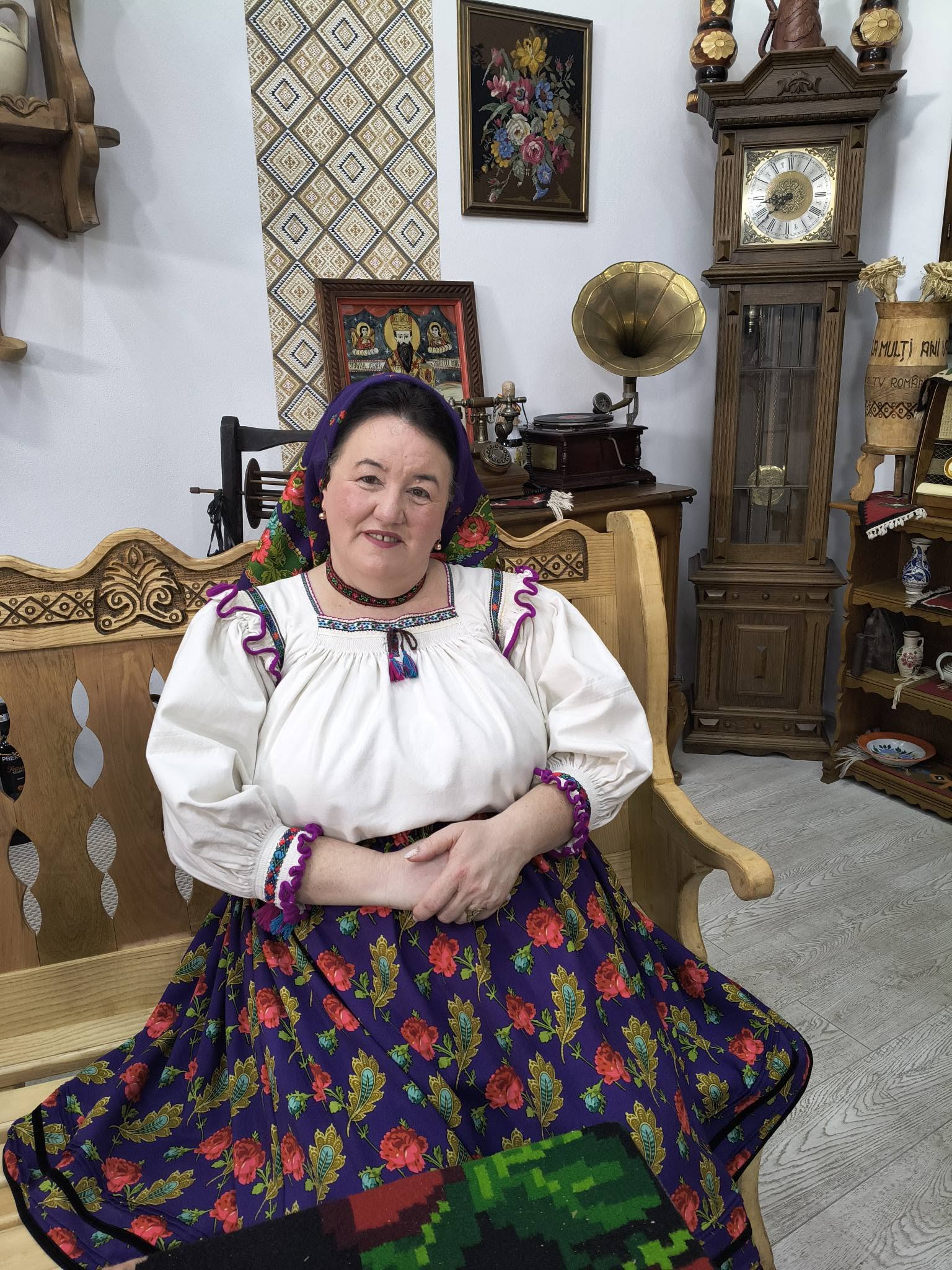 Ileana Matuș, fosta soție a lui Cristian Pomohaci, st&acirc;nd pe o bancă de lemn, &icirc;mbrăcată &icirc;n costum popular, st&acirc;nd cu m&acirc;inile &icirc;mpreunate