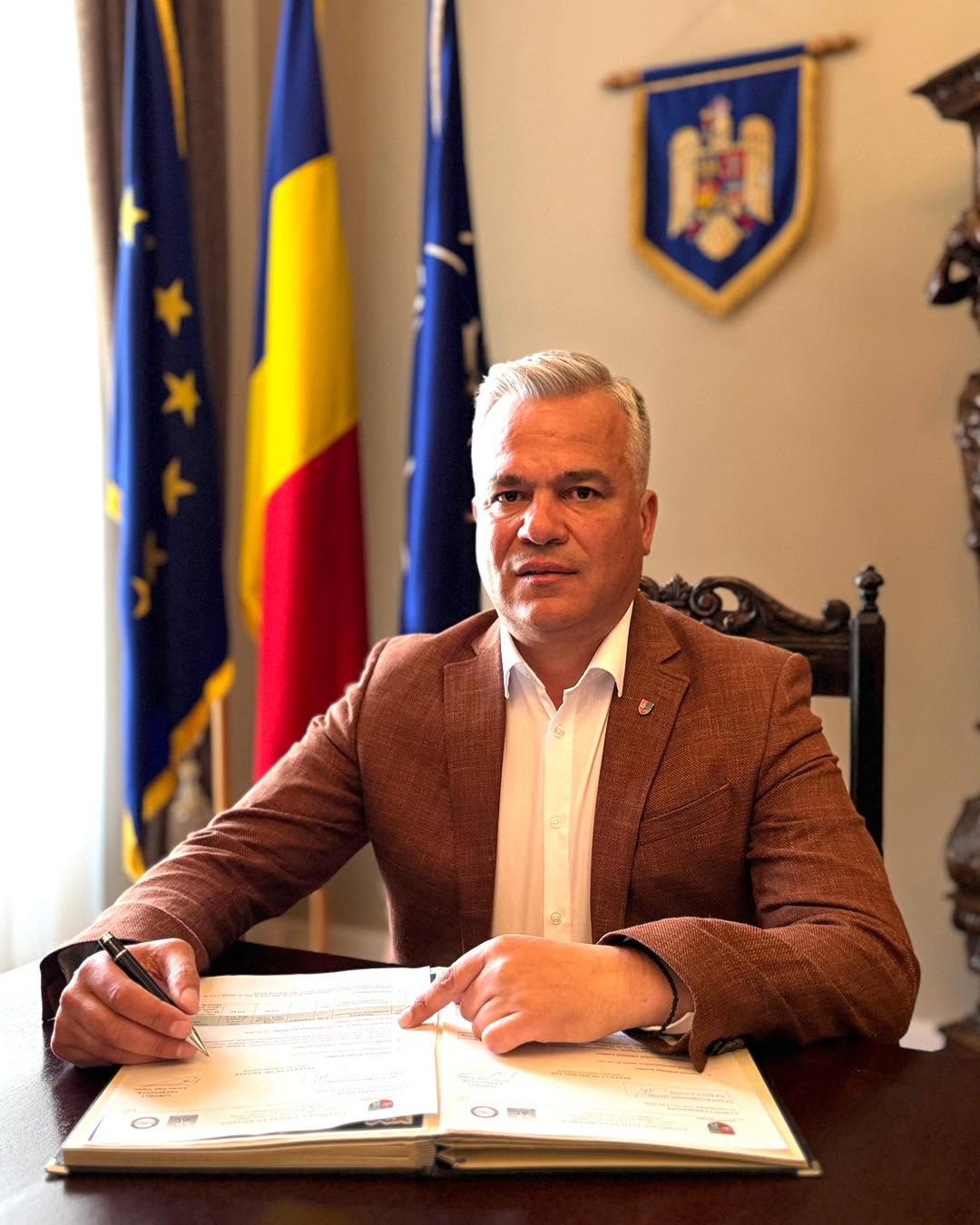 Adrian Veștea în sacou maro și cămașă albă, stând cu o mână pe un registru și ținând stiloul în cealaltă mână, arătând că este pregătit să semneze