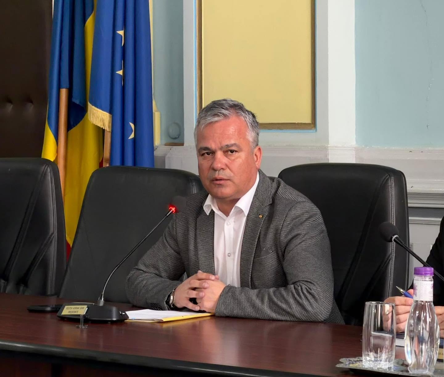 Adrian Veștea st&acirc;nd la birou, cu m&acirc;inile &icirc;mpreunate, &icirc;mbrăcat &icirc;ntr-un sacou gri și o cămașă albă