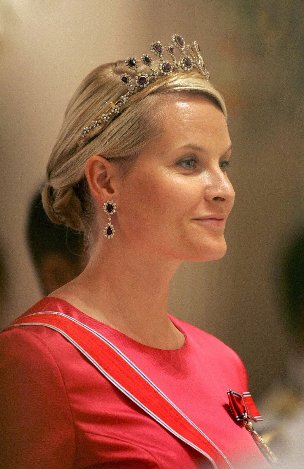 Mette-Marit, prințesa a Norvegiei &icirc;ntr-o rochie roz, cu părul prins &icirc;n coc, coroană pe cap și cercei lungi cu pietre prețioase