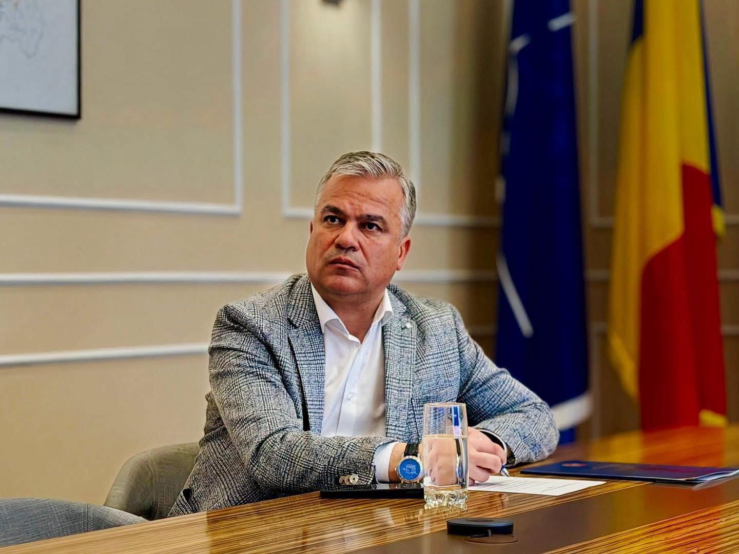Adrian Veștea &icirc;n birou, semn&acirc;nd un act, &icirc;mbrăcat &icirc;n sacou gri &icirc;n carouri și cămașă albă, purt&acirc;nd un ceas din piele la m&acirc;nă