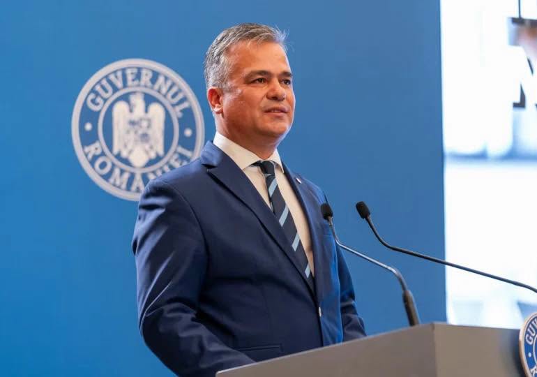 Adrian Veștea la Guvern, vorbind la microfon &icirc;n fața unei audiențe, &icirc;mbrăcat &icirc;n costum albastru și purt&acirc;nd cravată asortată