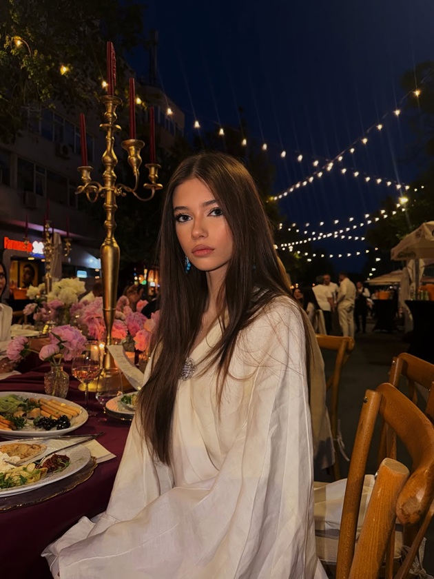 Mikaela Albu &icirc;ntr-o rochie albă cu m&acirc;neci largi și lungi, st&acirc;nd pe scaun la o masă festivă, cu m&acirc;ncarea &icirc;n față