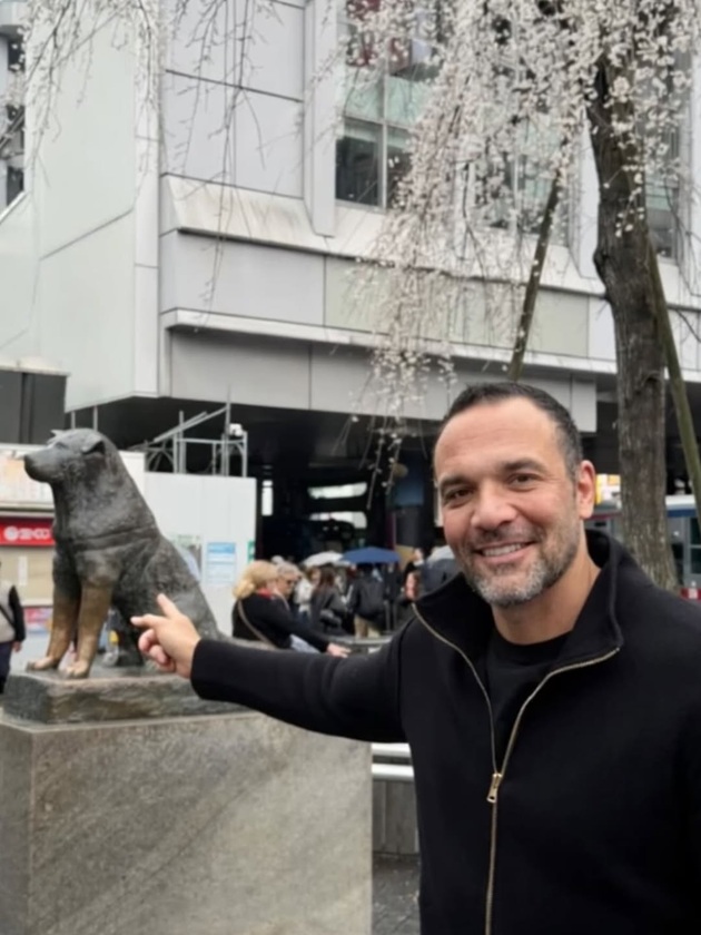 Alexandru Ion Țiriac z&acirc;mbitor, &icirc;mbrăcat &icirc;n negru, arăt&acirc;nd cu degetul spre o statuie &icirc;n formă de c&acirc;ine