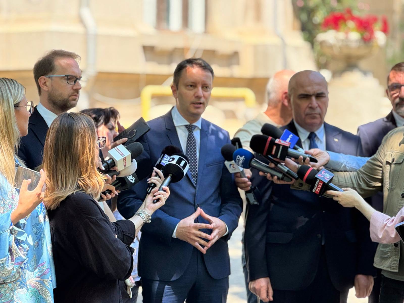 Siegfried Mureșan, &icirc;n costum albastru, cu cămașă albă și cravată cu picățele, țin&acirc;ndu-și m&acirc;inile &icirc;mpreunate, &icirc;n timp ce stătea l&acirc;ngă Ilie Bolojan și vorbea &icirc;n fața jurnaliștilor de la mai multe televiziuni