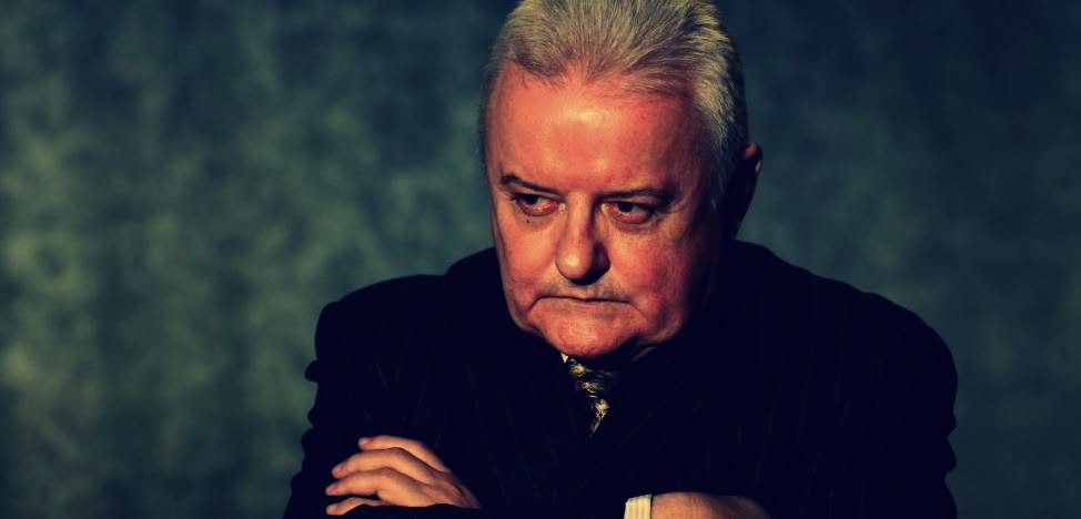 Irinel Columbeanu st&acirc;ns serios, privind &icirc;ntr-o parte, &icirc;n timp ce stătea l&acirc;ngă un perete gri, cu m&acirc;inile &icirc;ncrucișate, &icirc;mbrăcat &icirc;n costum negru
