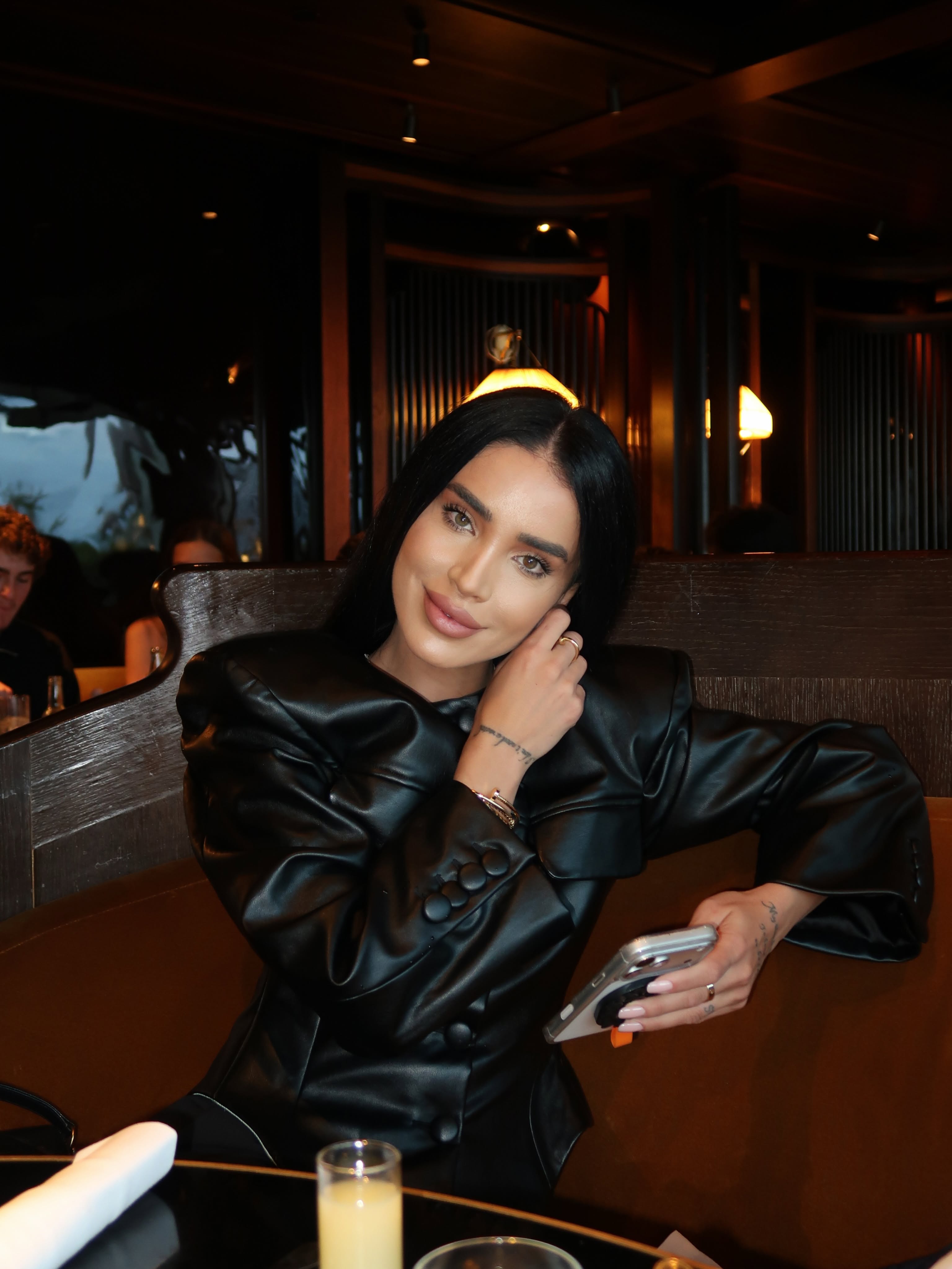 Adelina Pestrițu st&acirc;nd la masa unui restaurant, sprijinită de spătarul canapelei, cu telefonul &icirc;n m&acirc;nă, &icirc;n timp ce era &icirc;mbrăcată &icirc;ntr-o rochie de piele.