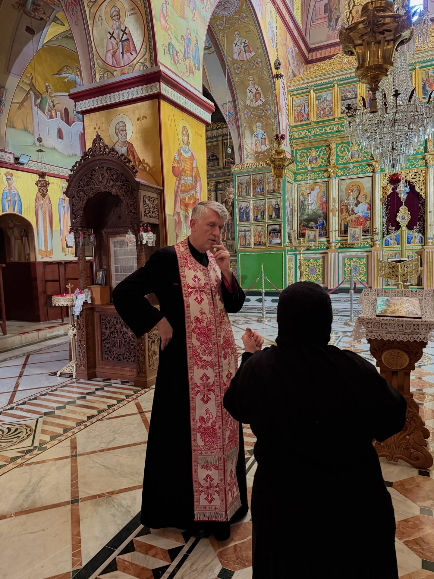 Cristian Pomohaci în biserică, îmbrăcat în haine preoțești, gânditor, stând de vorbă cu o maică, în timp ce ținea o mână în șold și una la bărbie