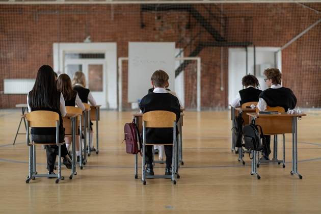 Elevi în uniformă, într-o sală de clasă spațioasă, așezați în bănci individuale, cu ghiozdanele agățate de marginea băncii