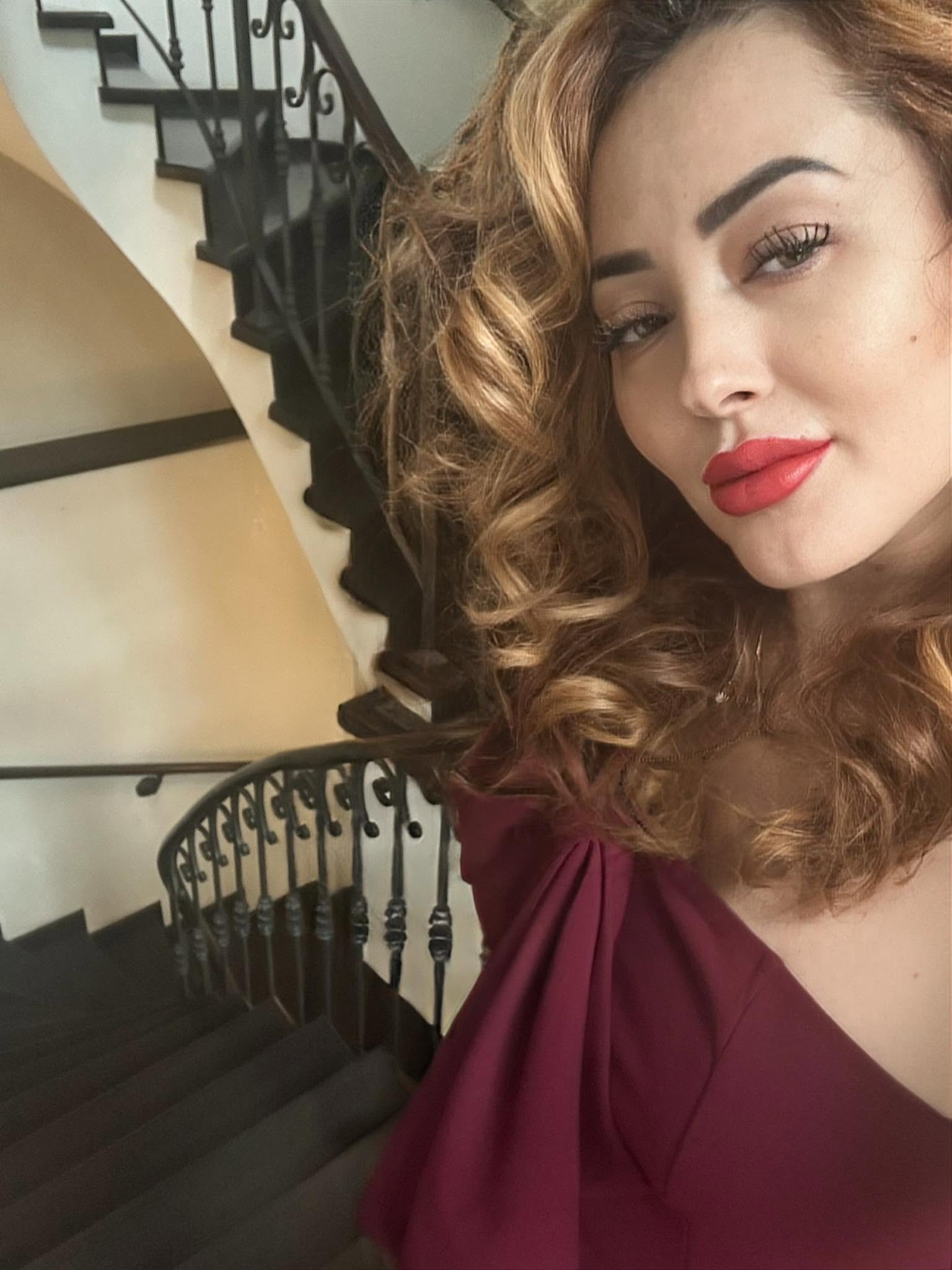 Nidia Moculrscu &icirc;ntr-o rochie roșie, cu părul ondulat, făc&acirc;ndu-și un selfie &icirc;n timp ce stătea sprijinită de balustrada unei scări