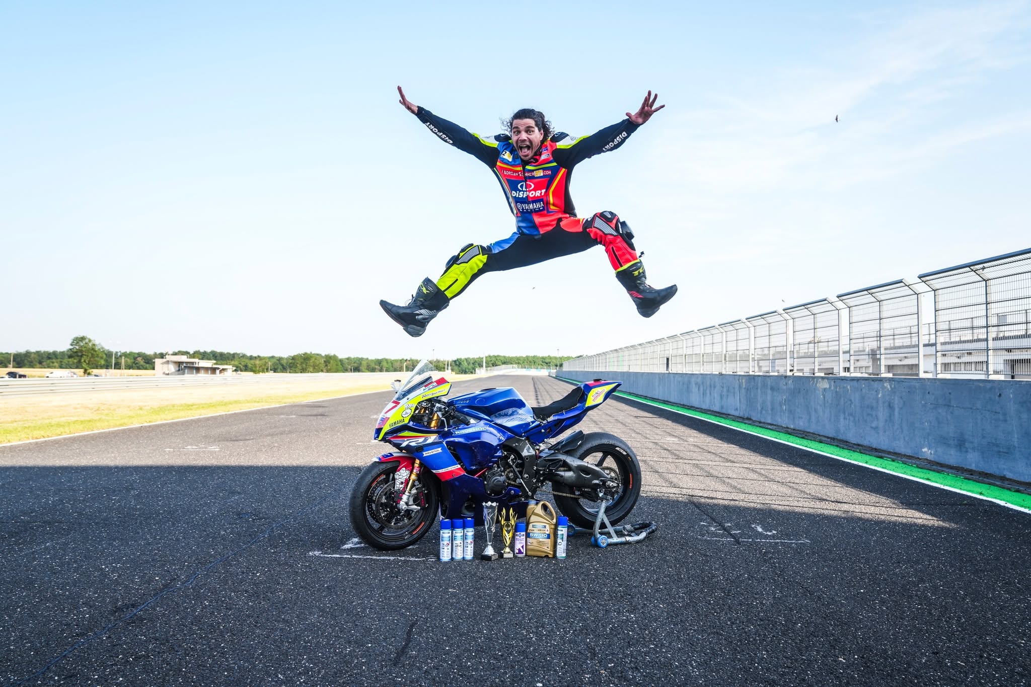 pilotul Adrian Rus execut&acirc;nd o săritură deasupra motocicletei sale l&acirc;ngă care se află și c&acirc;teva dintre trofeele sale