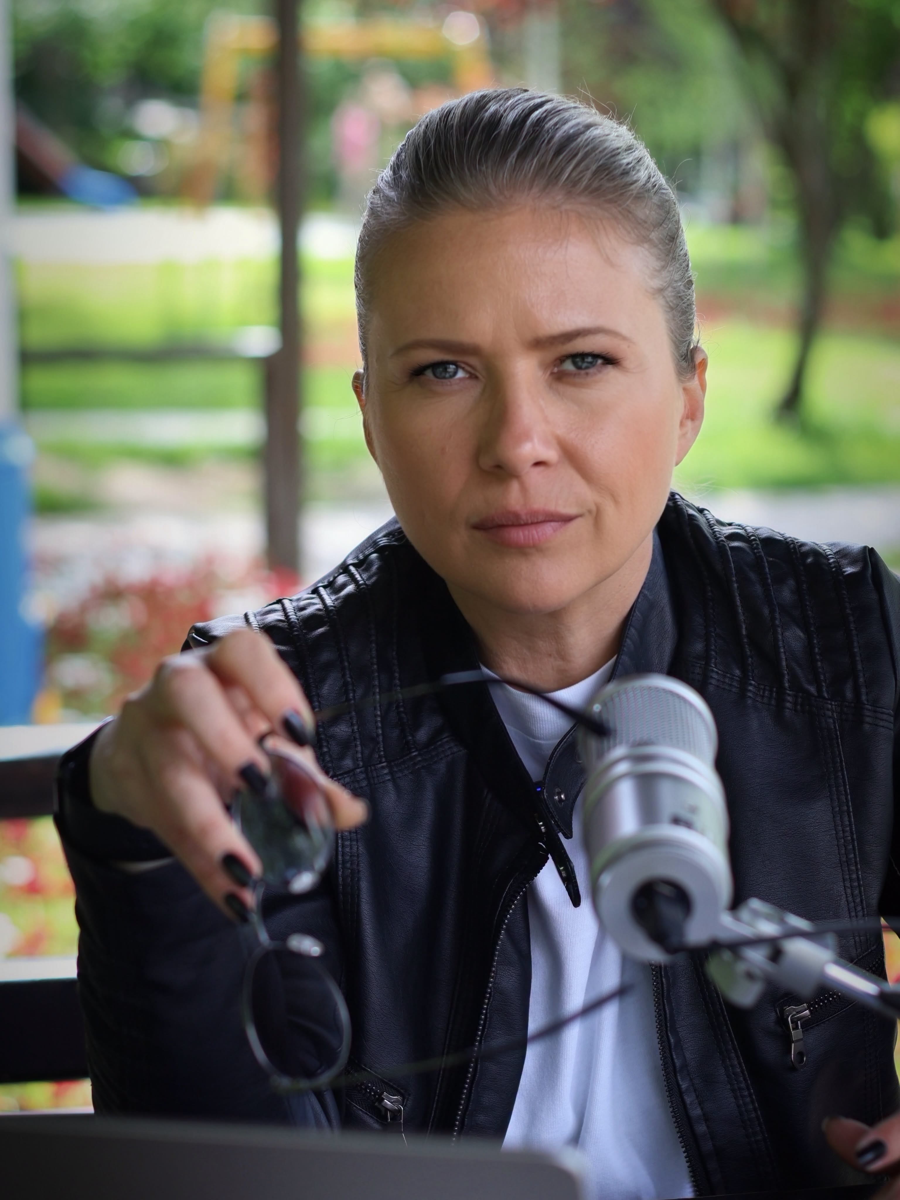 Sonia Simionov &icirc;n aer liber, pe o bancă de lemn, cu un microfon &icirc;n față, țin&acirc;nd o pereche de ochelari de soare &icirc;n m&acirc;nă și privind serioasă spre obiectivul camerei