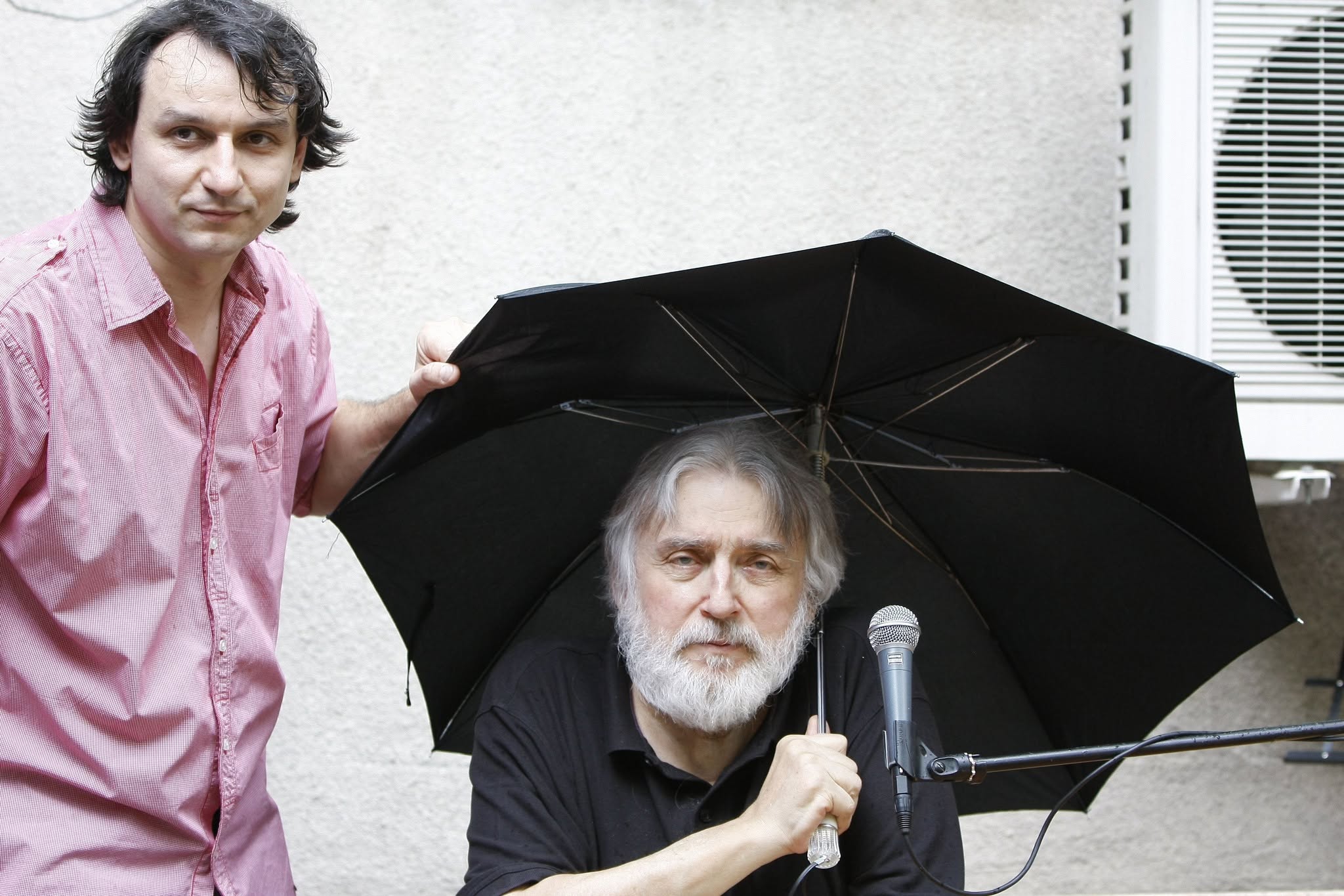 Adrian Păunescu la 67 de ani, st&acirc;nd la o masă, cu un microfon &icirc;n față și cu o umbrelă deasupra capului, alături de fiul său, Andrei Păunescu, care &icirc;i ține umbrela
