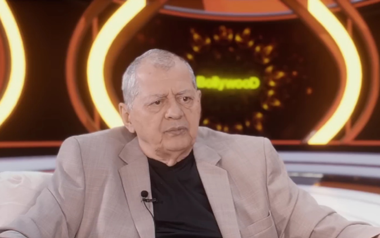 Octavian Ursulescu &icirc;ntr-un tricou negru și un sacou bej, cu umeri supradimensionați, st&acirc;nd pe o canapea, &icirc;ntr-un platou de televiziune