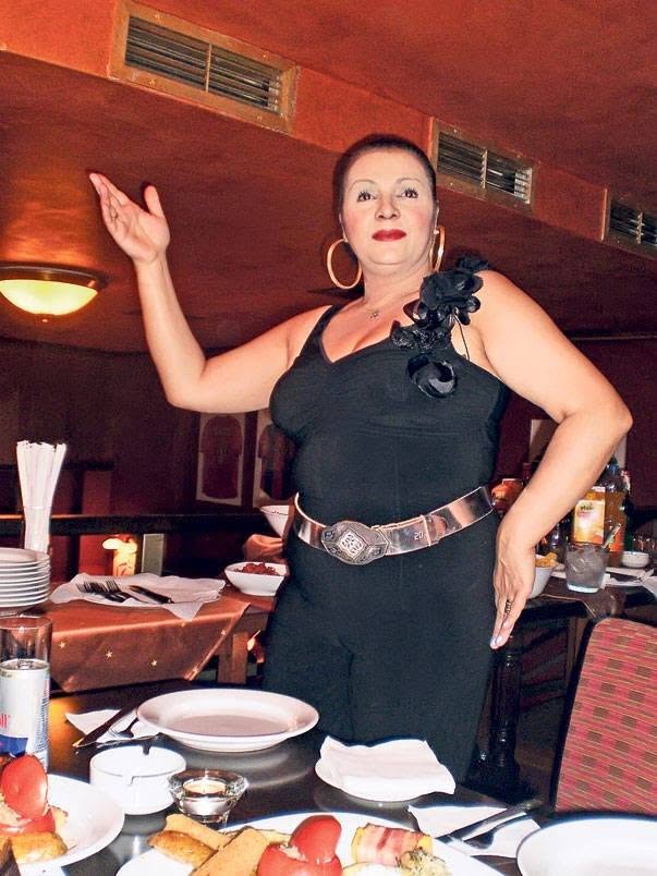 Cornelia Catanga z&acirc;mbitoare, &icirc;mbrăcată &icirc;ntr-o salopetă neagră, distr&acirc;ndu-se la o petrecere și dans&acirc;nd l&acirc;ngă masa plină cu m&acirc;ncare