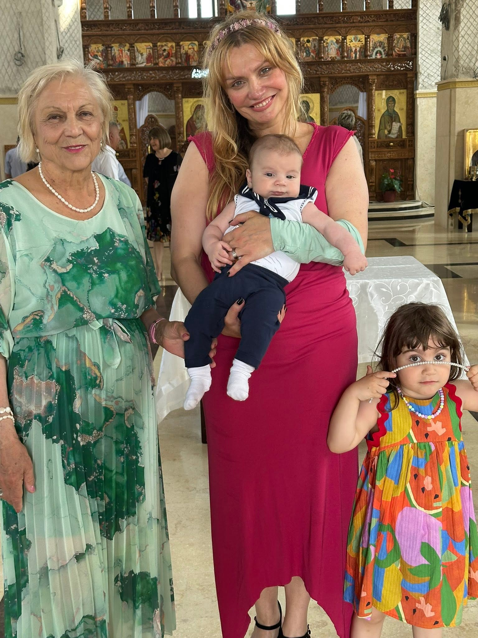 Cristina Cioran z&acirc;mbitoare &icirc;n biserică, alături de mama și copiii ei, &icirc;n timp ce &icirc;l ține pe fiul său &icirc;n brațe și poartă o rochie vișinie, mulată și lungă