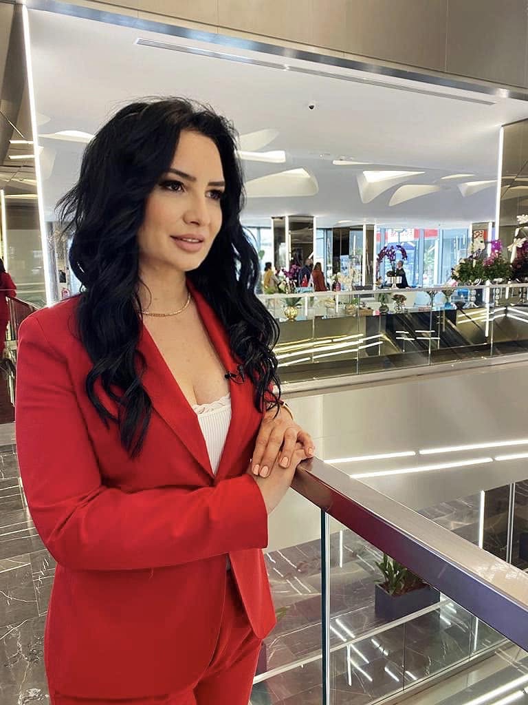 Eva Pavel &icirc;ntr-un costum roșu, cu părul ondulat, st&acirc;nd sprijinită de balustrada unei clădiri cu etaj