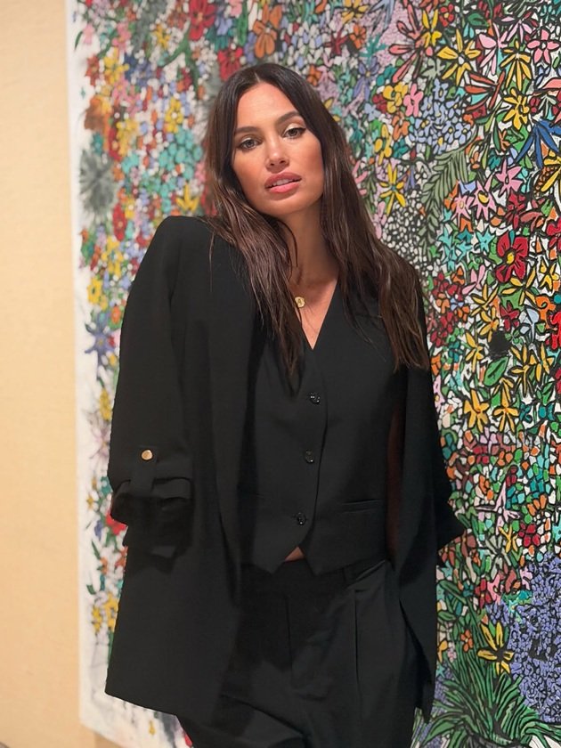 Alina&nbsp;Pușcău &icirc;mbrăcată complet &icirc;n negru, st&acirc;nd l&acirc;ngă un perete mozaic cu flori colorate, cu m&acirc;inile &icirc;n buzunarele de la pantaloni și cu sacoul pe umeri