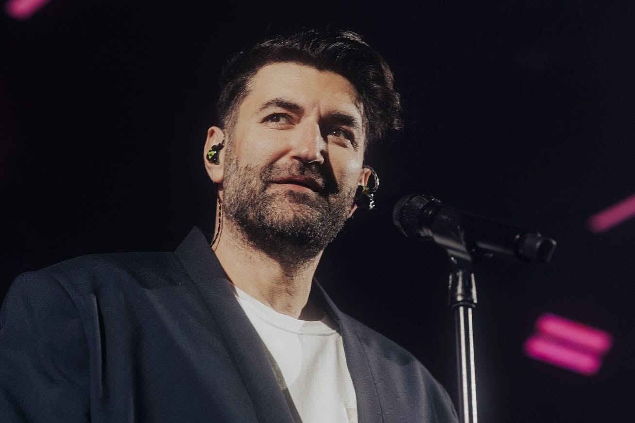 Smiley pe scenă, &icirc;n timpul unui concert, cu microfonul &icirc;n față, &icirc;mbrăcat &icirc;ntr-un tricou alb și un sacou negru, privind &icirc;ntr-o parte