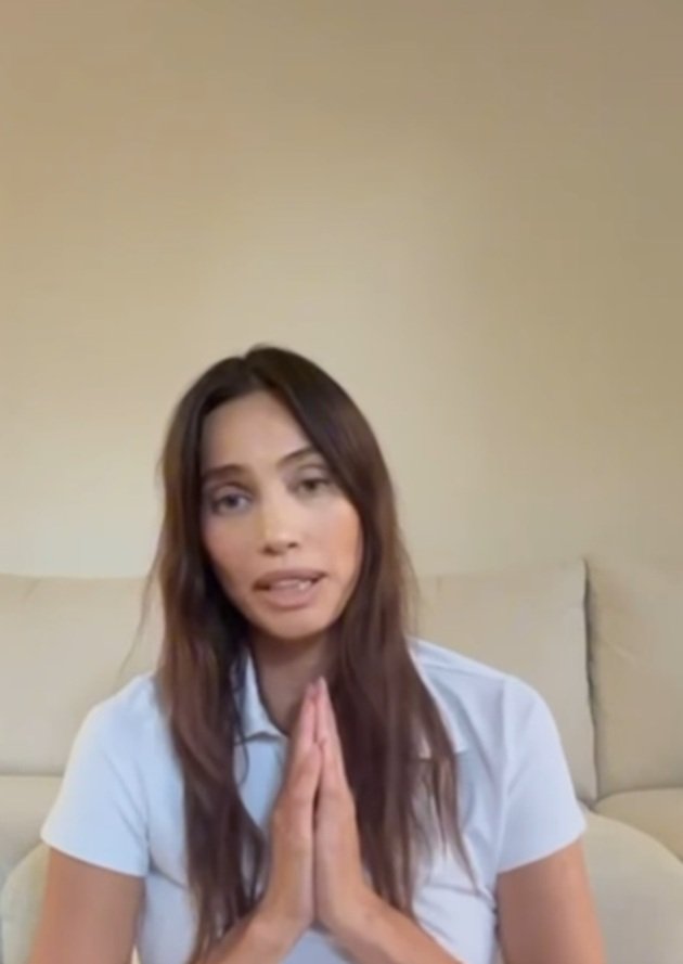 Alina Pușcău cu m&acirc;inile &icirc;mpreunate, rug&acirc;ndu-și fanii să se roage pentru ea, &icirc;n timp ce stătea pe jos, &icirc;n fața unei canapele crem și purta un tricou alb cu guler
