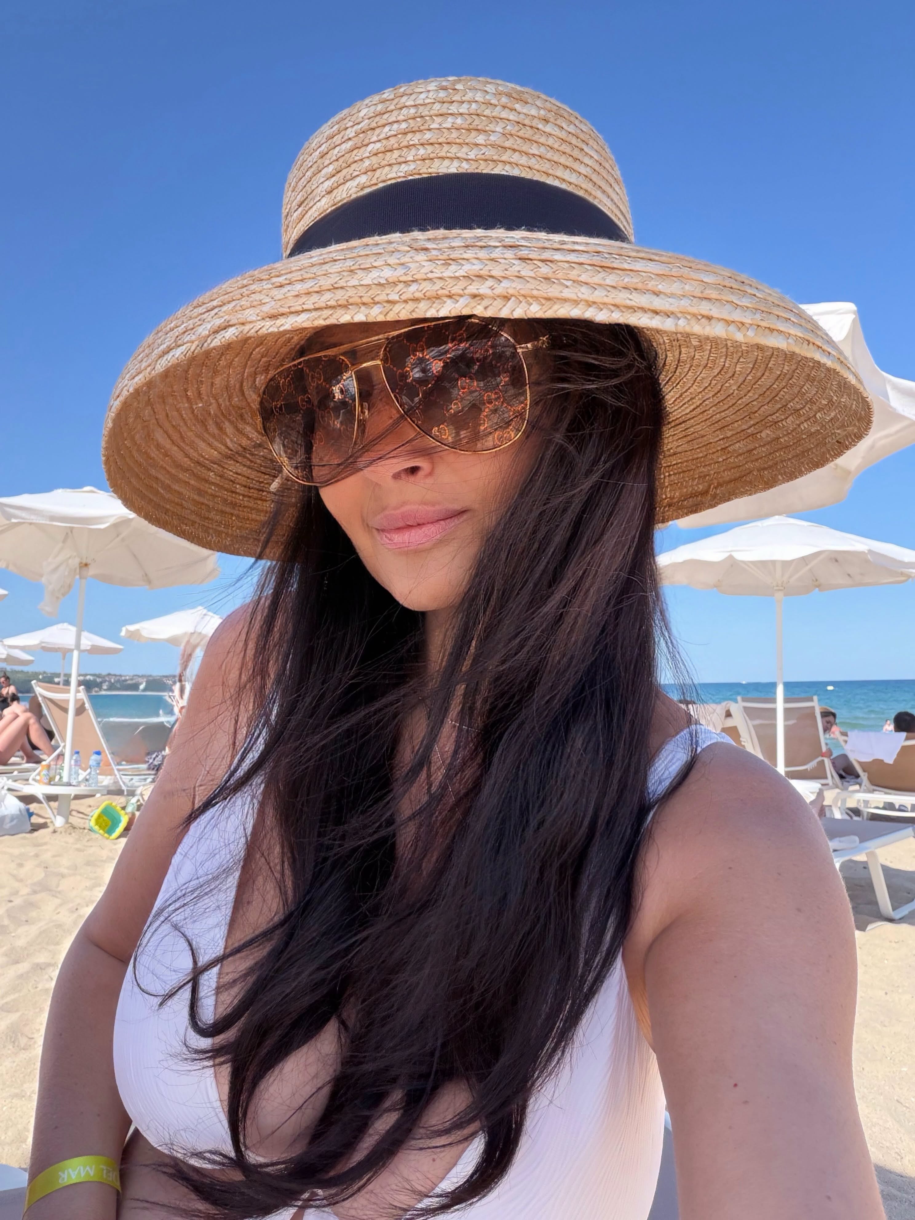 Gabriela Cristea la plajă, cu părul lung, desfăcut, cu pălărie pe cap și ochelari de soare la ochi, &icirc;n timp ce era &icirc;mbrăcată &icirc;n costum de baie alb