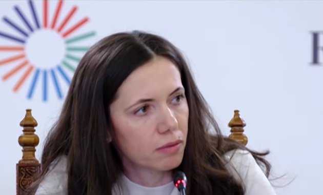 Mirabela Grădinaru st&acirc;nd pe scaun de lemn, cu spătar &icirc;nalt, &icirc;n timpul unei conferințe publice, &icirc;n timp ce era &icirc;mbrăcată &icirc;n alb și avea părul dat după urechi