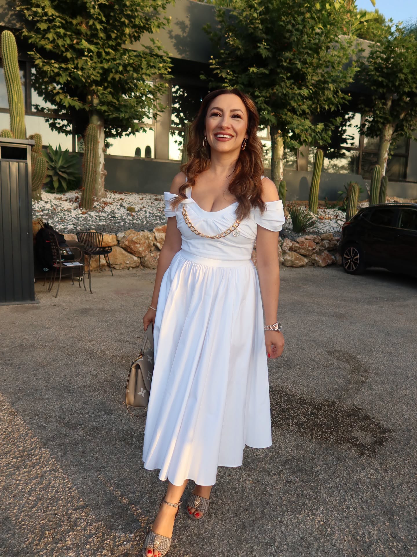 Andra Măruță într-o rochie albă, care are un detaliu auriu în zona pieptului, ținând în mână o geantă asortată cu sandalele pe care le avea în picioare
