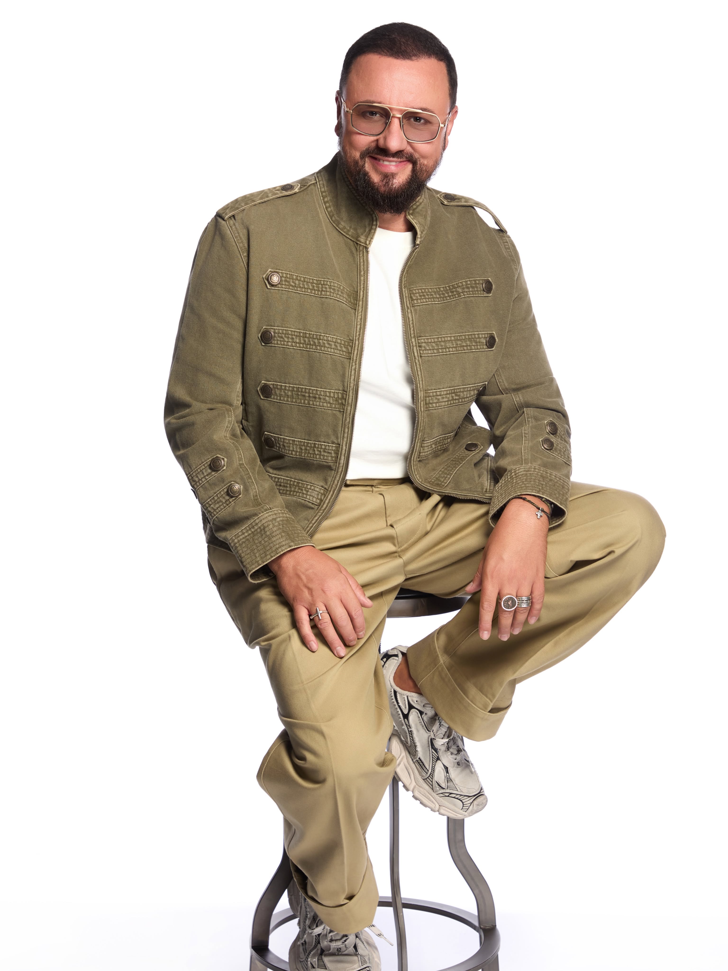 Cătălin Măruță &icirc;ntr-un studio foto, &icirc;n fața unui perete alb, st&acirc;nd pe un scaun, &icirc;mbrăcat &icirc;n haine bej și kaki, z&acirc;mbitor, cu ochelari de soare la ochi și cu inele pe degete