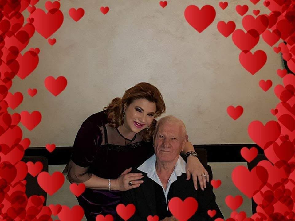 Mihaela Borcea z&acirc;mbitoare, &icirc;ntr-o rochie neagră, &icirc;mbrățiș&acirc;ndu-și tatăl care stătea pe scaun, &icirc;mbrăcat &icirc;n cămașă albă și pulover negru