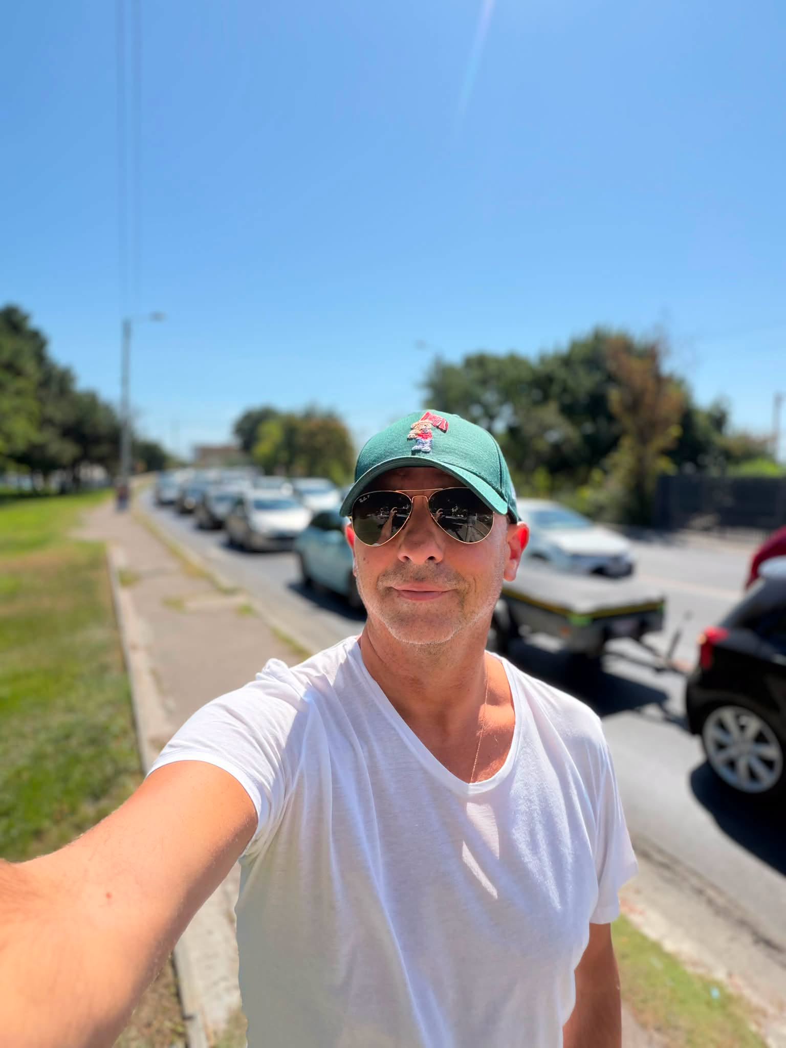 Dan Negru &icirc;mbrăcat &icirc;ntr-un tricou alb, cu șapcă verde pe cap și ochelari de soare la ochi, st&acirc;nd pe un trotuar, făc&acirc;ndu-și un selfie l&acirc;ngă mai multe mașini parcate pe marginea străzii