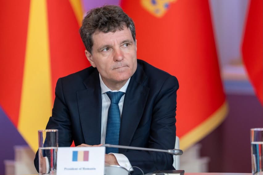 Nicușor Dan st&acirc;nd &icirc;n fața unei audiențe, cu microfonul l&acirc;ngă el și cu un pahar pe masă așezat l&acirc;ngă plăcuța cu funcția lui, &icirc;n timp ce &icirc;n spate se aflau mai multe steaguri.