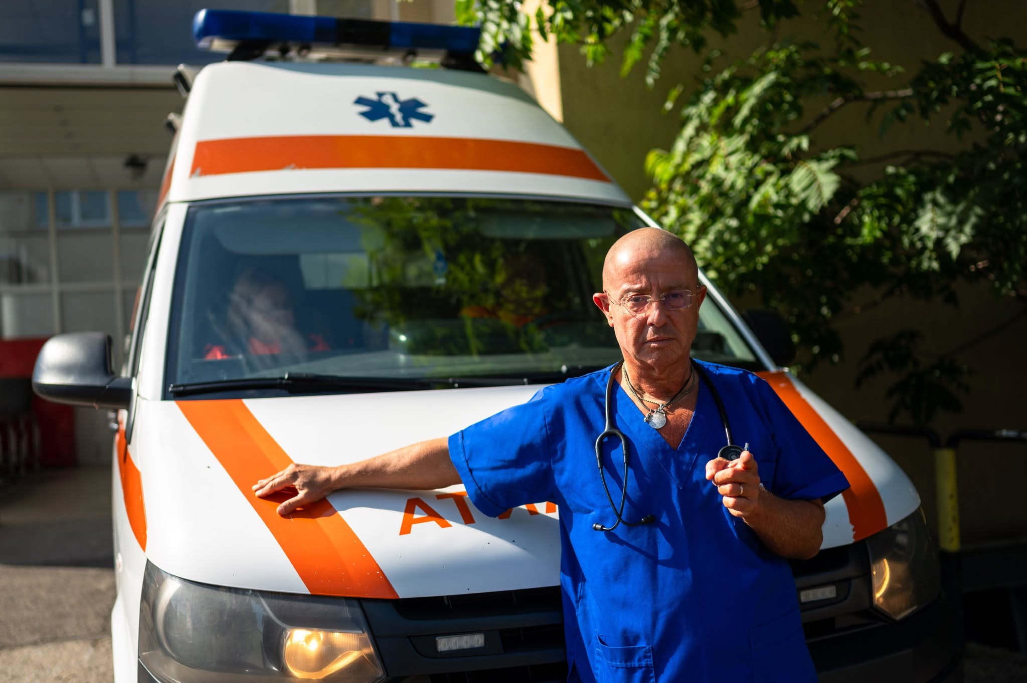 medicul&nbsp;Tudor&nbsp;Ciuhodaru st&acirc;nd sprijinit de o ambulanță, &icirc;mbrăcat &icirc;n hainele de serviciu și arăt&acirc;nd cu degetul spre obiectivul aparatului de fotografiat, &icirc;n timp ce purta la g&acirc;t un stetoscop și ochelari de vedere la ochi