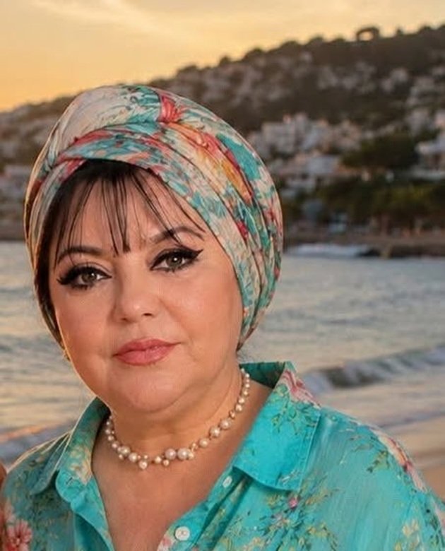 Actrița&nbsp;Corina Căpăț&acirc;nă purt&acirc;nd o cămașă turcoaz cu flori, un turban asortat și mărgele la g&acirc;t
