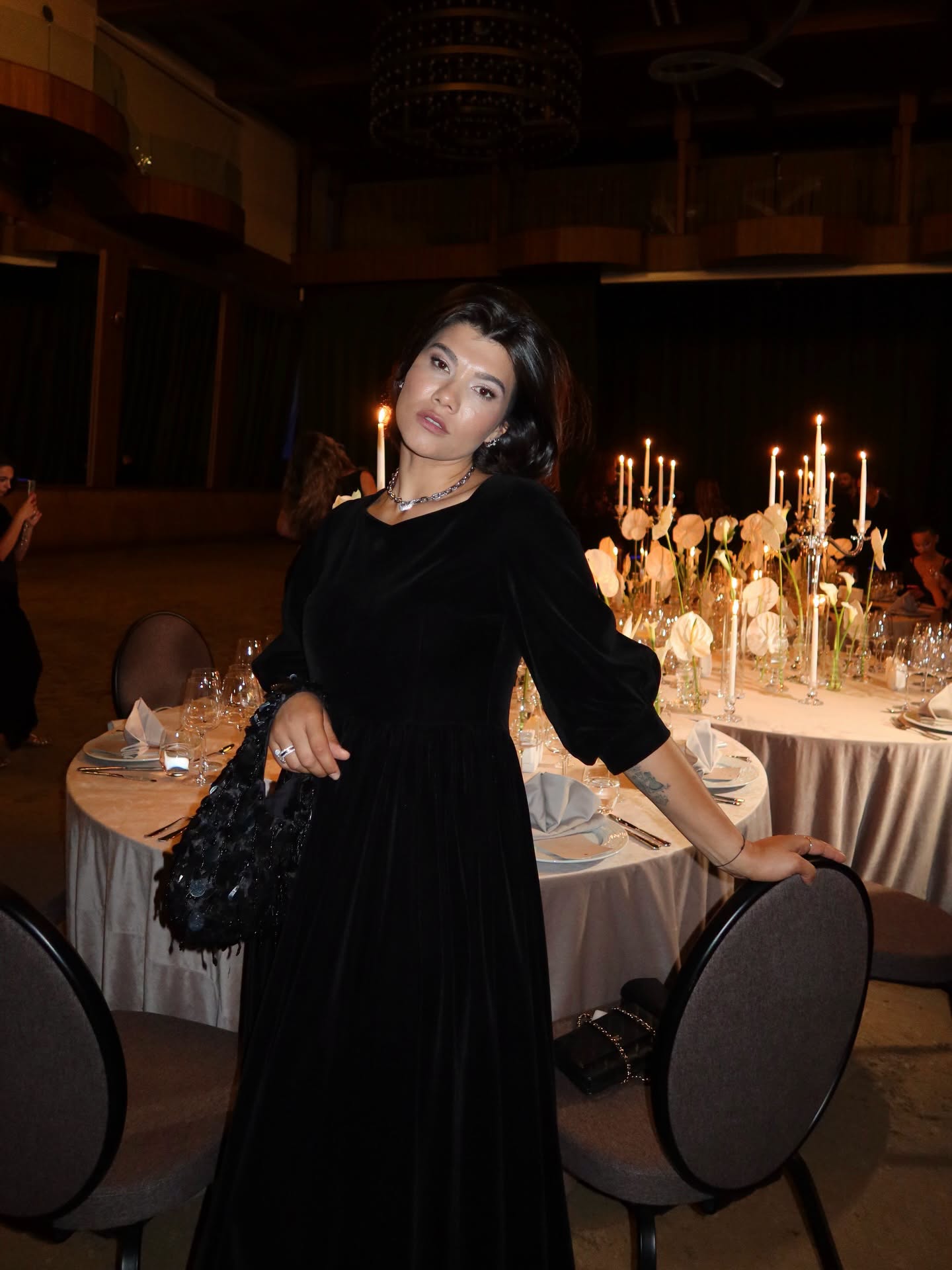 Ileana Sterp la un eveniment de seară, îmbrăcată în rochie neagră lungă, cu o geantă cu paete pe mână, stând lângă mai multe mese vestive, cu lumânări aprinse pe ele