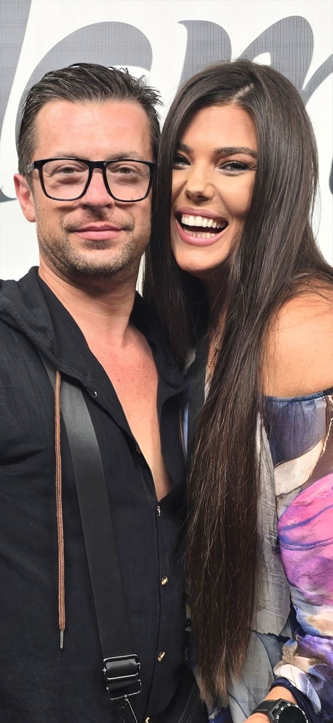 Paula Seling z&acirc;mbitoare &icirc;n brațele lui Alexandru Gorgos, &icirc;mbrăcată &icirc;ntr-o rochie cu flori, &icirc;n timp ce el purta o cămașă neagră și ochelari de vedere