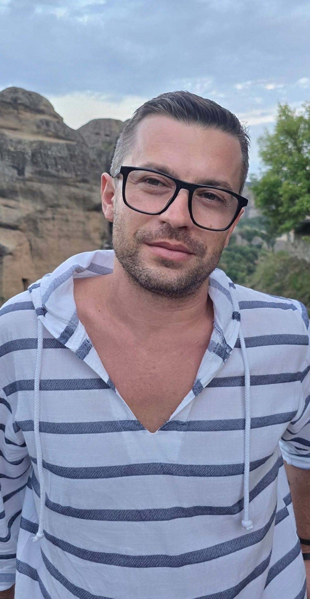 Alexandru&nbsp;Gorgos &icirc;ntr-o bluză albă cu dungi și guler &icirc;n V, purt&acirc;nd ochelari de vedere