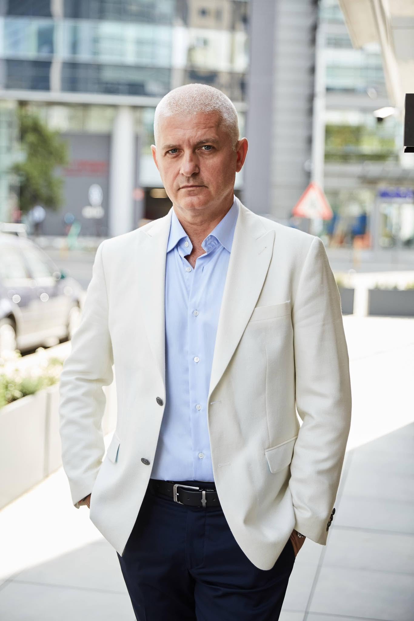 Virgil Ianțu &icirc;mbrăcat &icirc;n pantaloni negri, cu sacou alb și cămașă albă, st&acirc;nd cu m&acirc;inile &icirc;n buzunar, privind serios spre obiectivul camerei