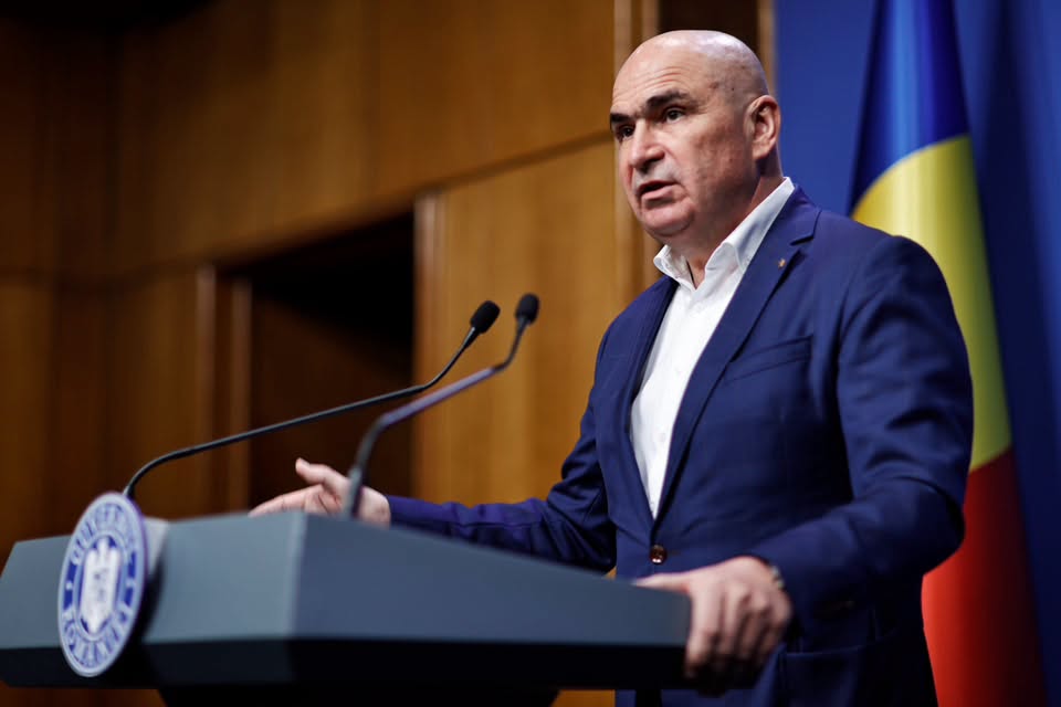 Ilie Bolojan la pupitrul cu sigla Guvernului, cu două microfoane &icirc;n față, &icirc;mbrăcat &icirc;n sacou bleumarin și cămașă albă