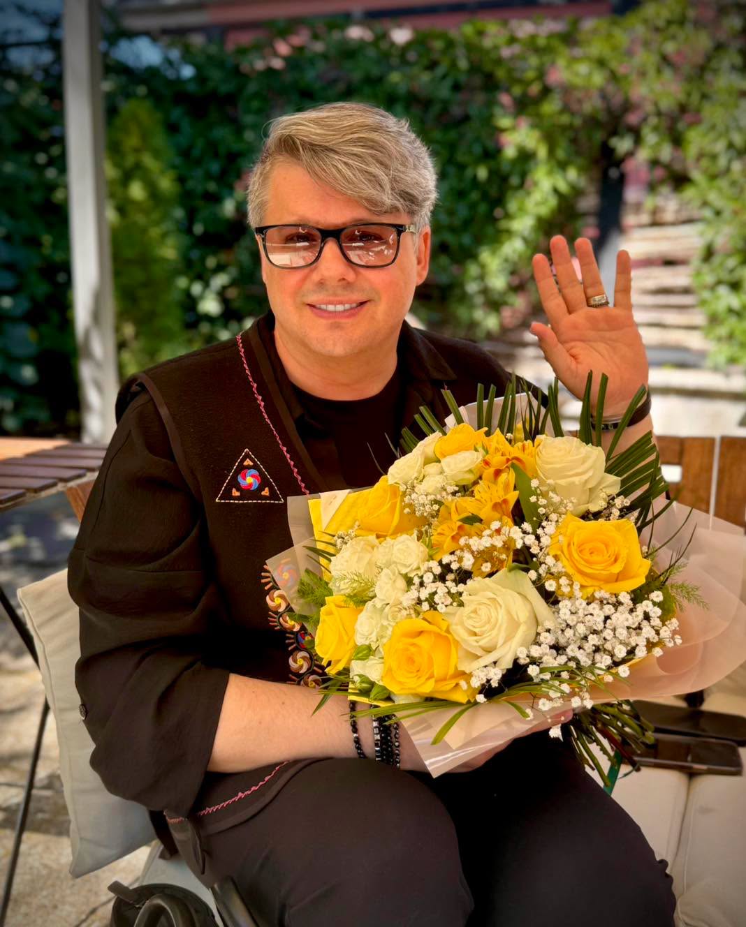 Fuego st&acirc;nd pe scaun la o terasă și țin&acirc;nd &icirc;n m&acirc;nă un buchet de trandafiri galbeni și albi cu gypsophila