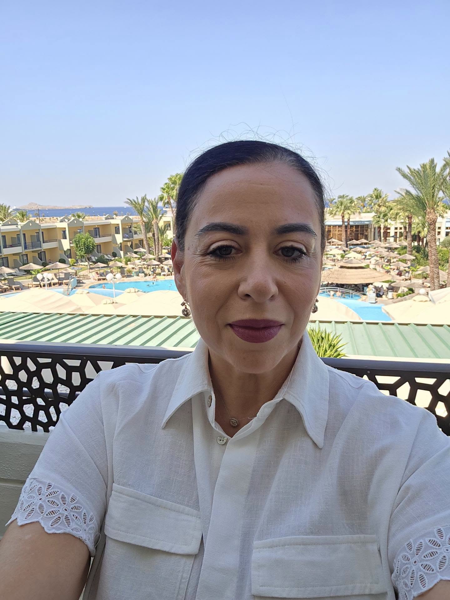 Oana Roman &icirc;n cămașă albă, făc&acirc;ndu-și un selfie pe balconul camerei de hotel cu vedere la piscina &icirc;nconjurată de palmieri