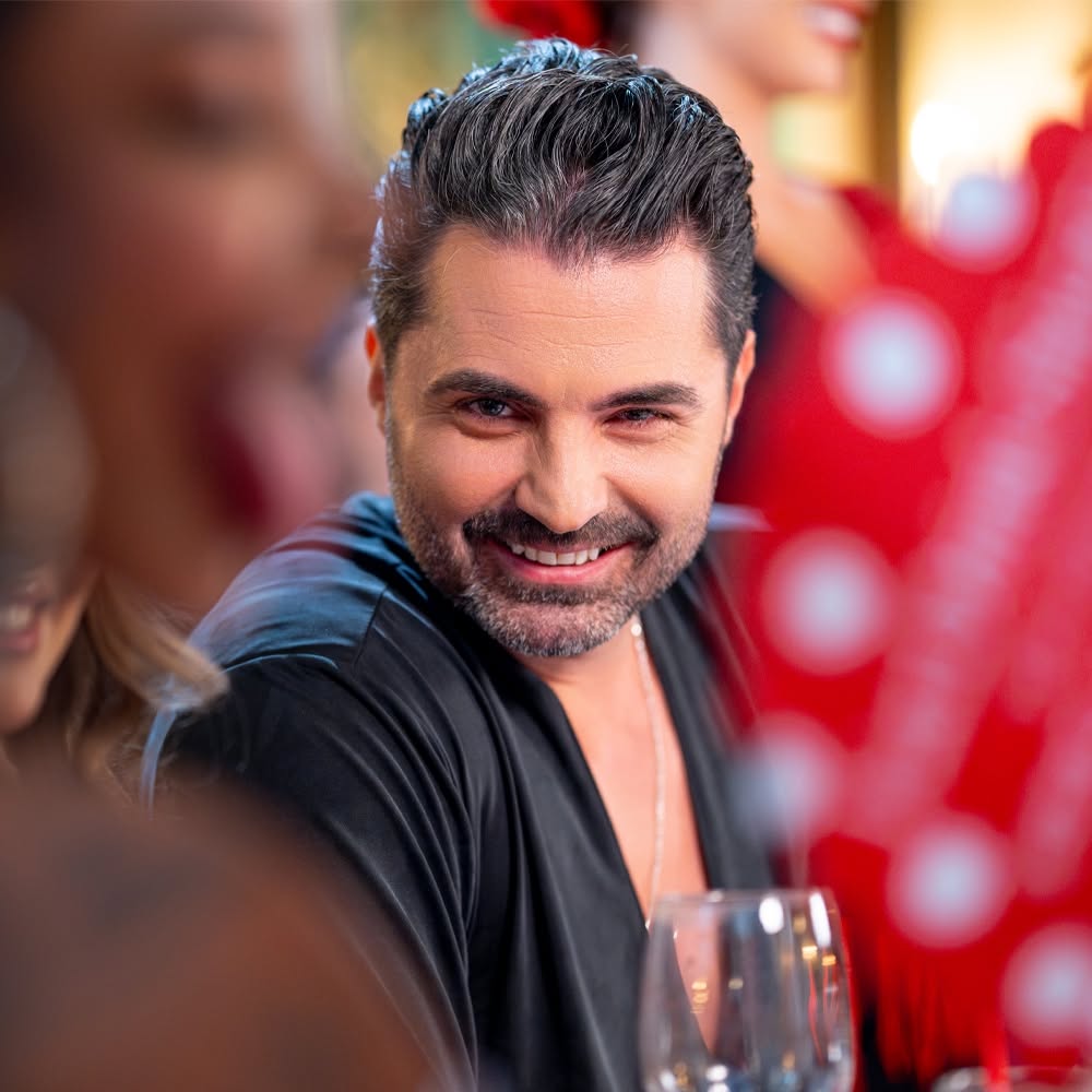 Pepe z&acirc;mbitor, st&acirc;nd la o masă, cu niște pahare &icirc;n față, purt&acirc;nd un tricou negru decoltat și un lanț la g&acirc;t