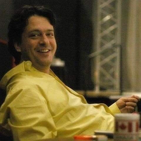 Regizorul Alexandru Darie, fiul lui Iurie Darie, z&acirc;mbitor, st&acirc;nd relaxat pe un scaun, &icirc;mbrăcat &icirc;ntr-o cămașă galbenă