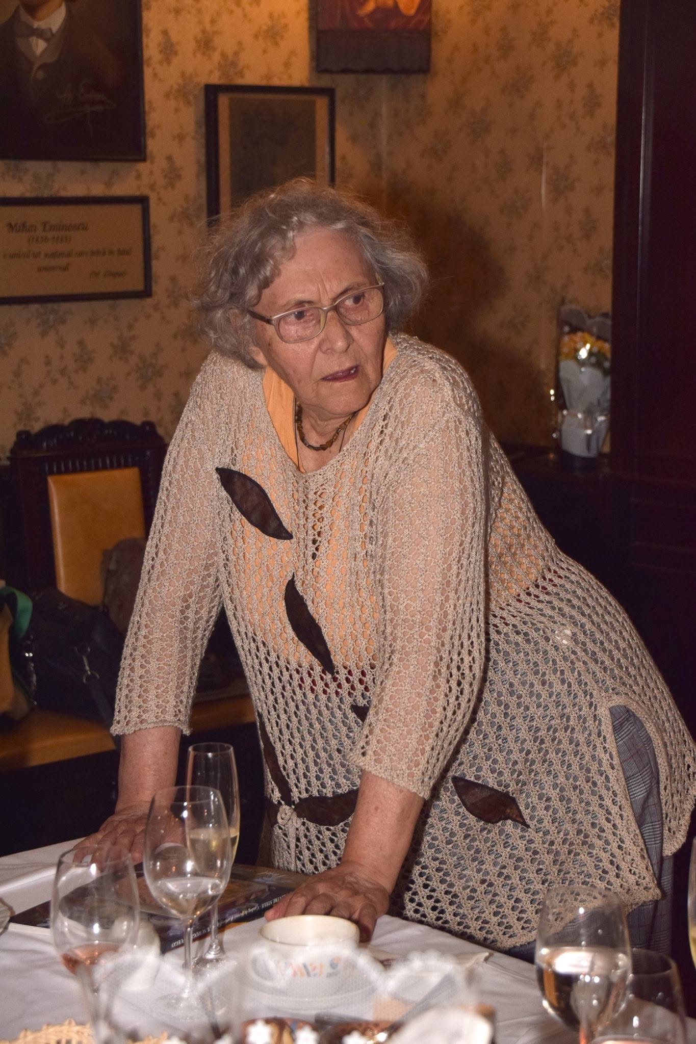 Actrița Adriana Piteșteanu sprijinindu-se de o masă, îmbrăcată cu un pulover lung stil plasă, purtând ochelari de vedere și privind într-o parte