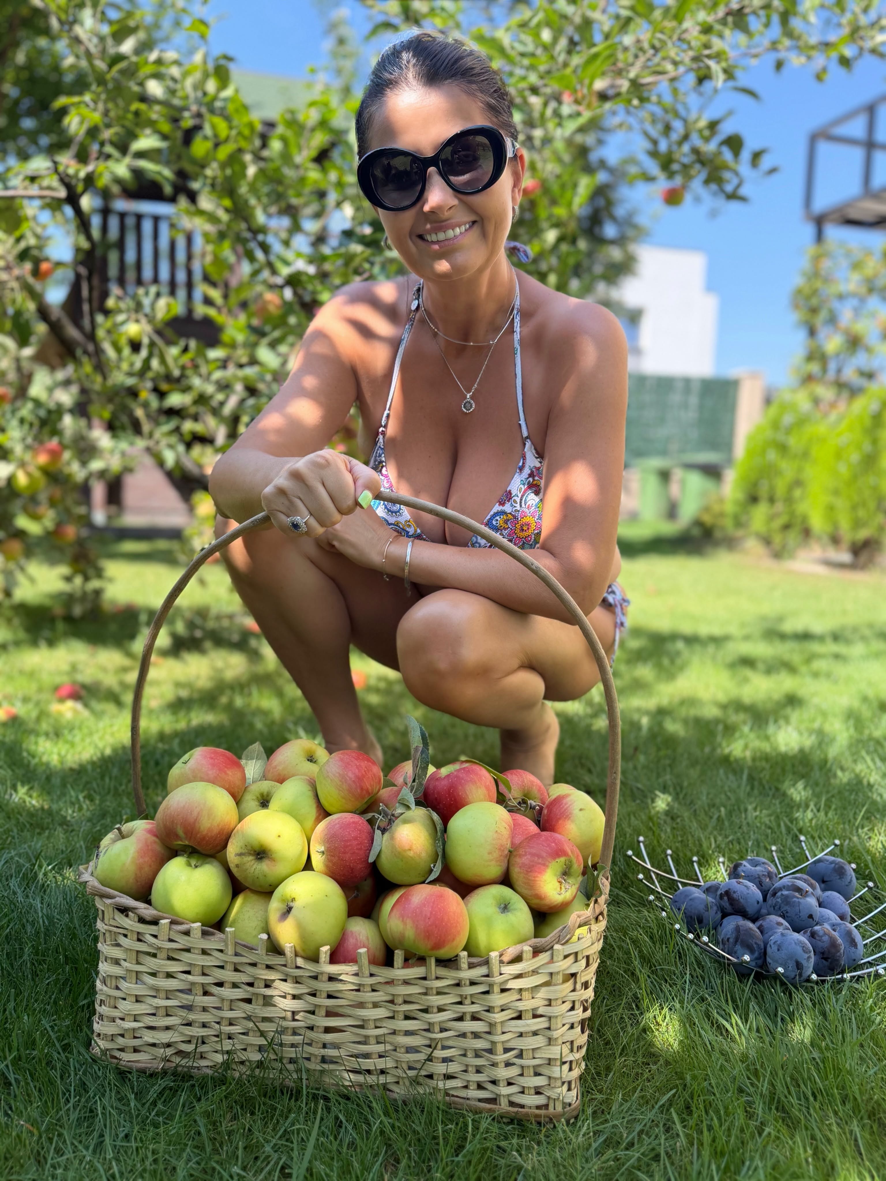 Gabriela Cristea stând pe vine în curtea casei sale, în spatele unor coșuri cu mere și prune culese de ea, în timp ce purta doar un costum de baie și ochelari de soare