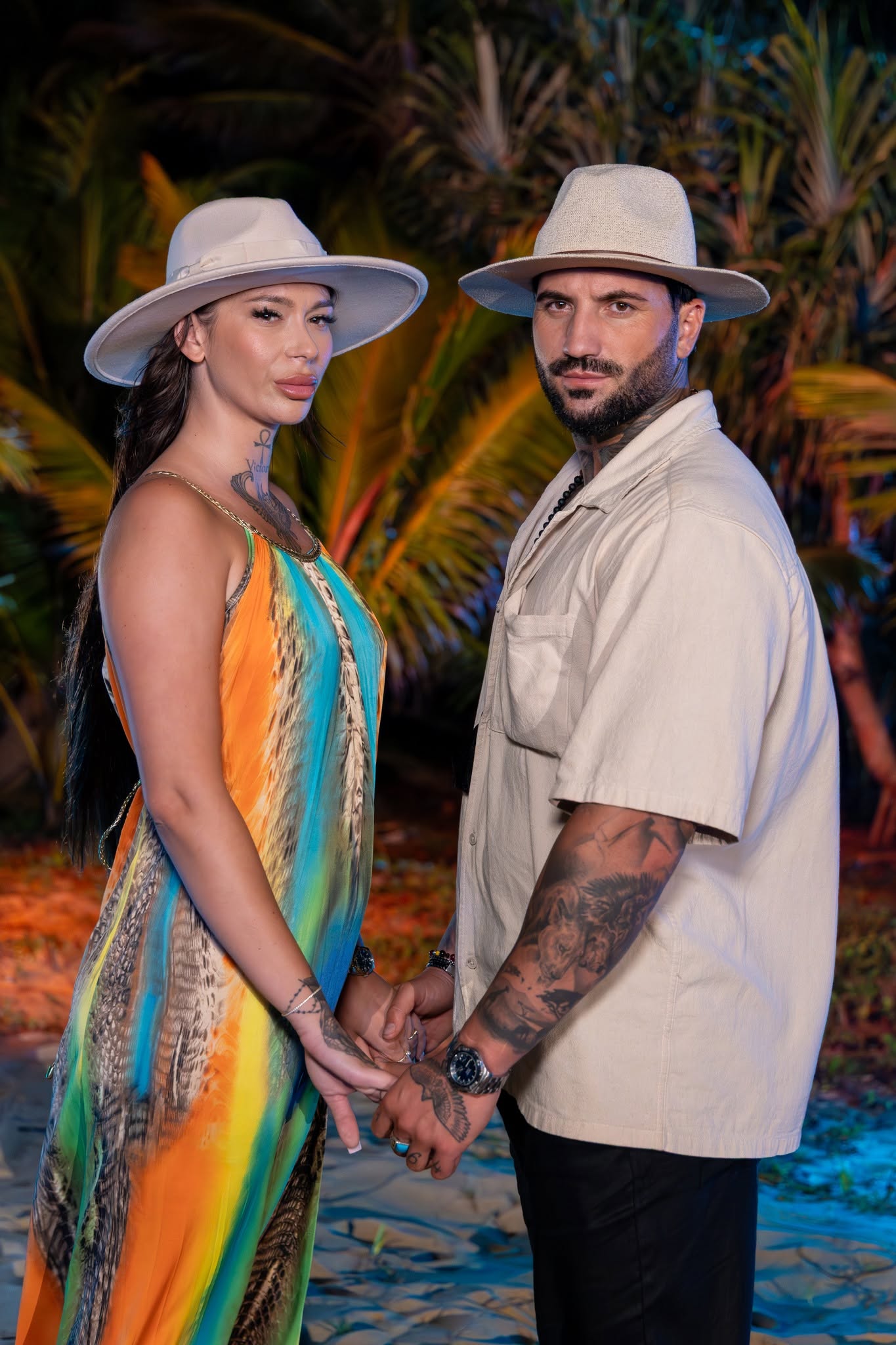 Armina și Marcel Andrei st&acirc;nd față &icirc;n față pe plajă, țin&acirc;ndu-se de m&acirc;ini, cu pălării pe cap, ea &icirc;mbrăcată &icirc;n rochie colorată și el &icirc;n pantaloni negri și cămașă bej
