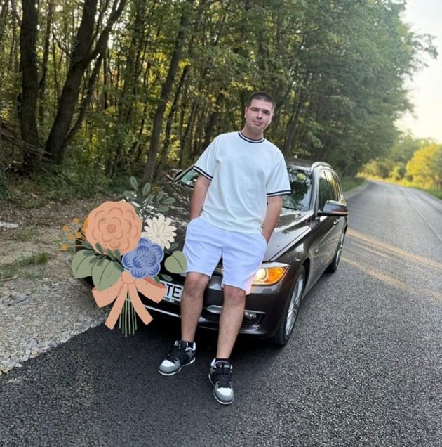 Riccardo&nbsp;Valentin Moraru, rom&acirc;nul de 20 de ani mort &icirc;n accidentul de muncă din Italia, &icirc;mbrăcat &icirc;n alb, st&acirc;nd cu m&acirc;inile &icirc;n buzunar, sprijinindu-se de o mașină neagră, la marginea unei păduri