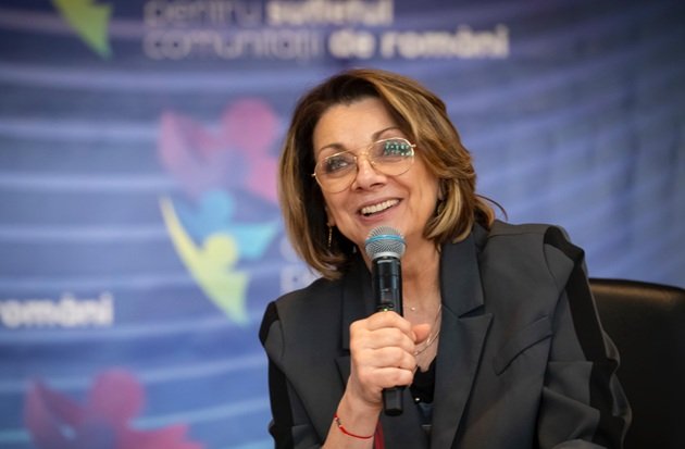 Carmen Tănase z&acirc;mbitoare, cu microfonul &icirc;n m&acirc;nă &icirc;n timpul unui eveniment public, &icirc;mbrăcată &icirc;ntr-un sacou gri &icirc;nchis cu detalii negre