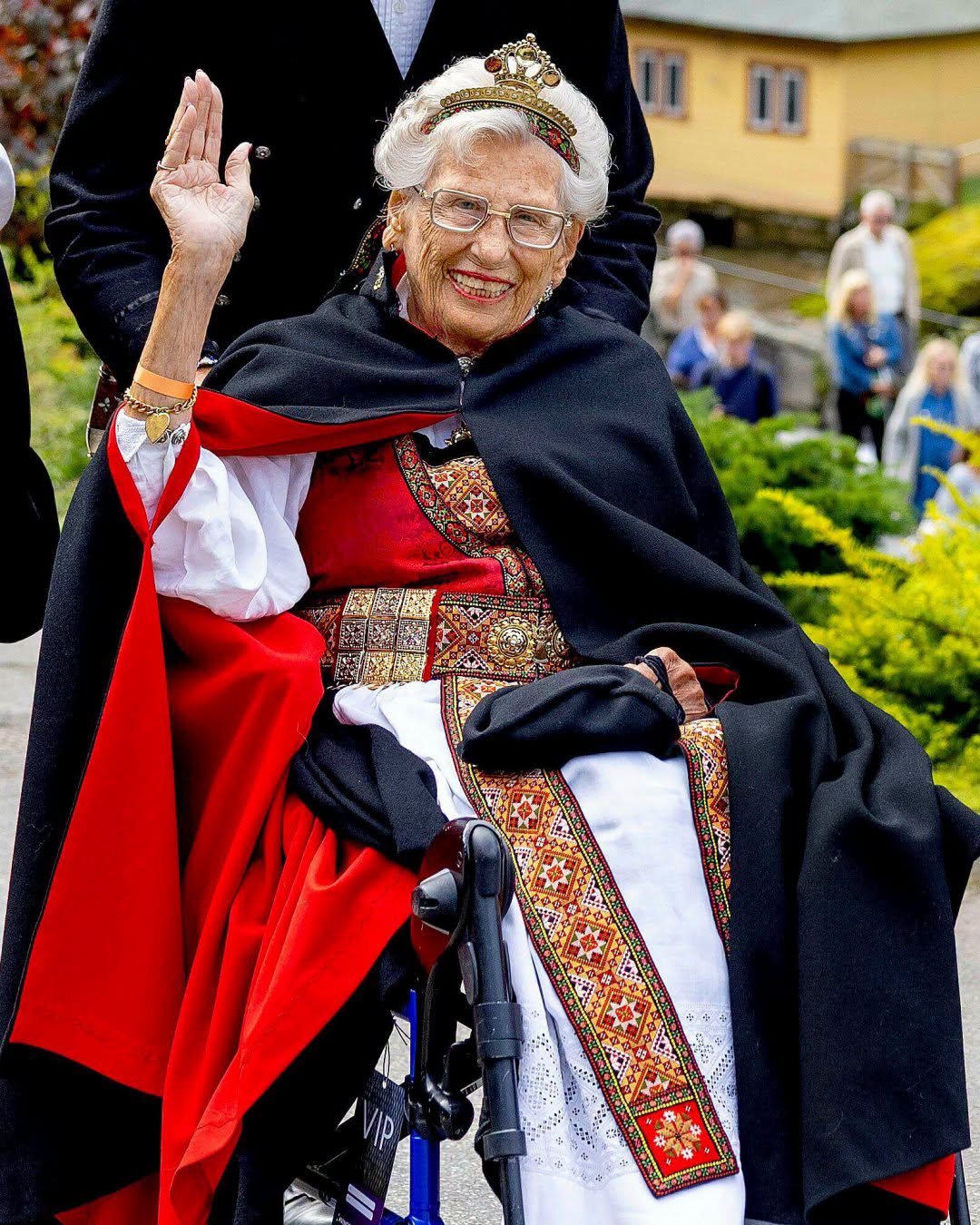 Prințesa&nbsp;Astrid&nbsp;a Norvegiei &icirc;mbrăcată &icirc;ntr-un costum tradițional, st&acirc;nd &icirc;n scaun cu rotile și făc&acirc;nd cu m&acirc;na, z&acirc;mbitoare, cu ochelari de vedere la ochi și coroană pe cap