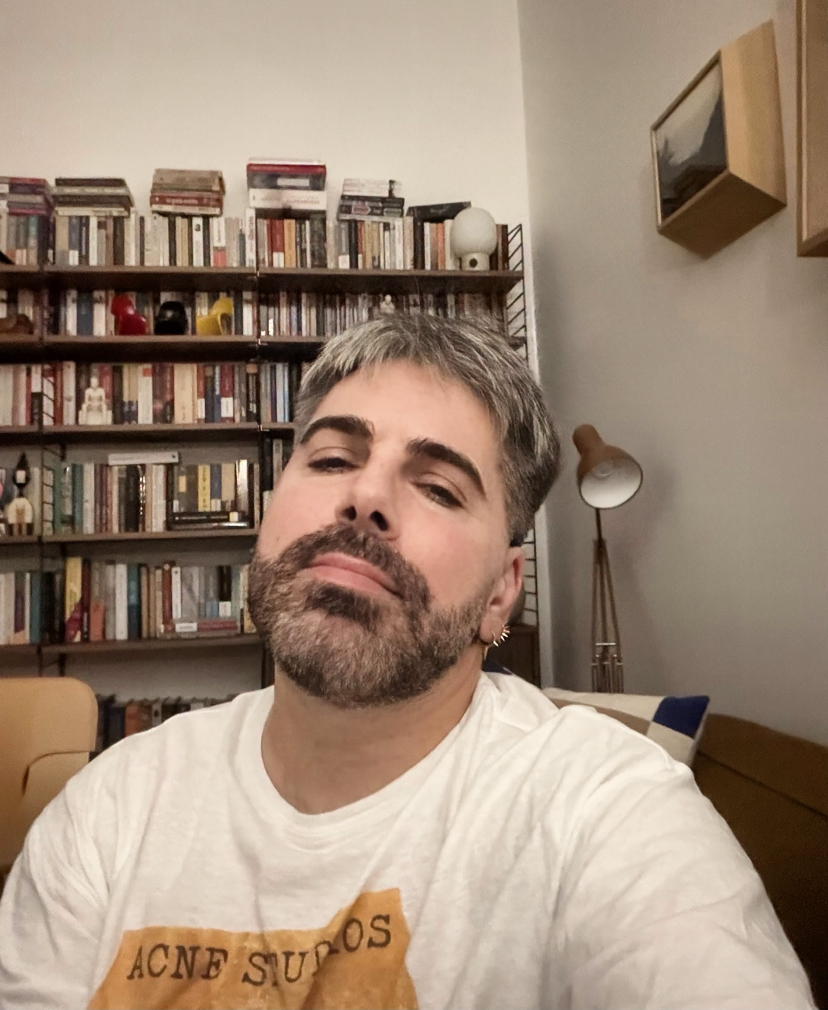 Maurice Munteanu &icirc;ntr-un tricou alb, st&acirc;nd &icirc;n casă, pe o canapea, &icirc;n fața unei biblioteci pline cu cărți și bibelouri