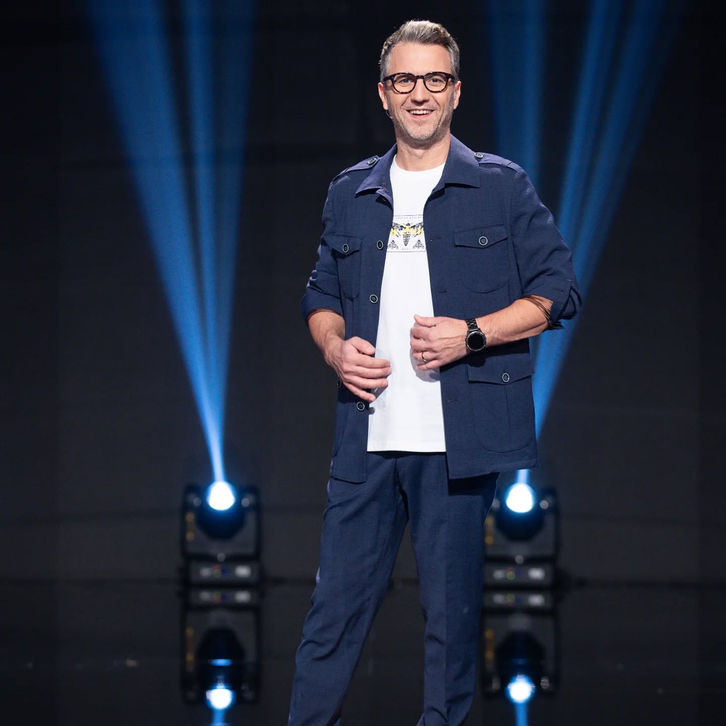 Andi Moisescu pe scenă, &icirc;n lumina reflectoarelor, &icirc;ntr-un costum casual bleumarin, pe sub care are un tricou alb